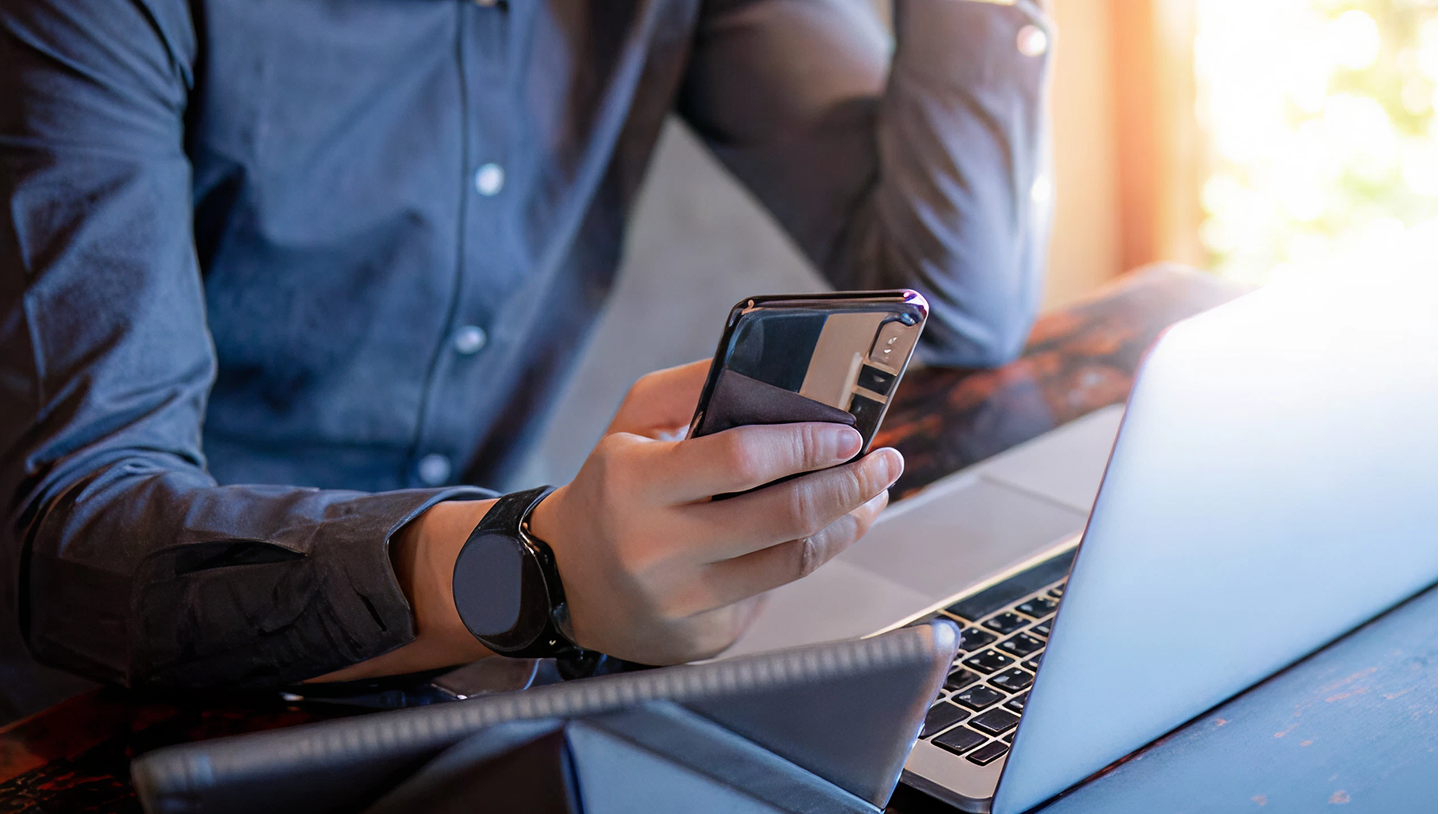 man holding a smartphone while sitting in front of his laptop